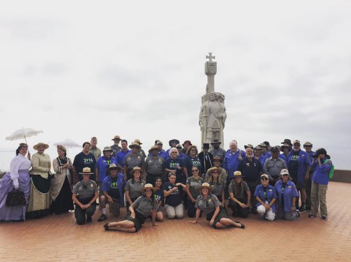 A group photo of Cabrillo National Monument staff and volunteers, some in uniform and some in period clothing, standing in front of the Cabrillo statue on an overcast day.