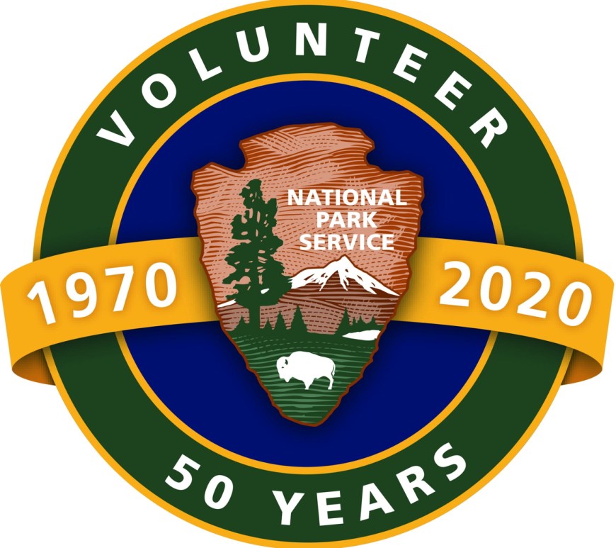 A circular logo of the National Park Service’s Volunteers-In-Parks (VIP) 50th anniversary. In the middle, a yellow banner with the years 1970 and 2020 in white font are separated by the National Park Service Arrowhead symbol in front a blue circle. A thin, yellow boundary separates the blue circle and a thicker green rim that contains the words “Volunteer” and “50 years” on the top and bottom, respectively.