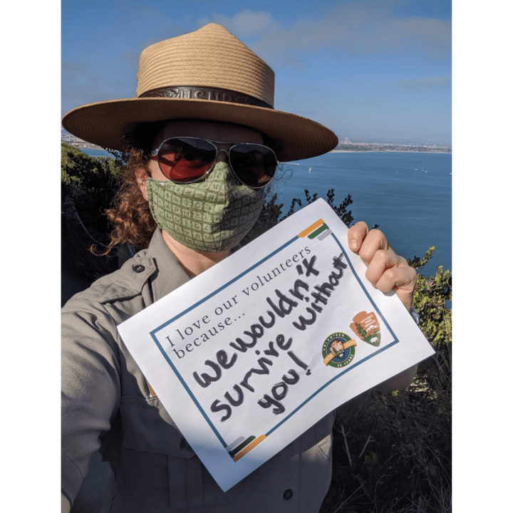 An up-close woman with a Park Ranger uniform in a green mask and sunglasses stands outside in front of the San Diego Bay holds up a sign that says, “I love our volunteers because…we wouldn’t survive without you!”