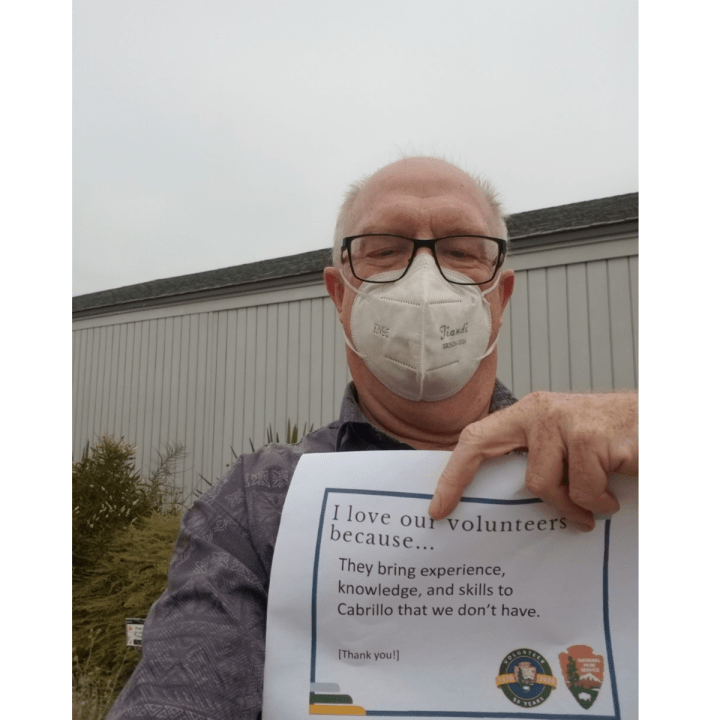 An up-close man with glasses and a white mask stands in front of a white building and some green shrubs and holds up a sign that says, “I love our volunteers because…they bring experience, knowledge, and skills to Cabrillo that we don’t have. Thank you!”