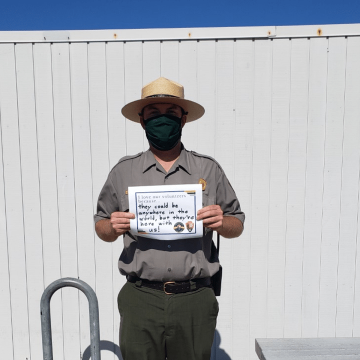 An up-close man with glasses and a white mask stands in front of a white building and some green shrubs and holds up a sign that says, “I love our volunteers because…they bring experience, knowledge, and skills to Cabrillo that we don’t have. Thank you!”