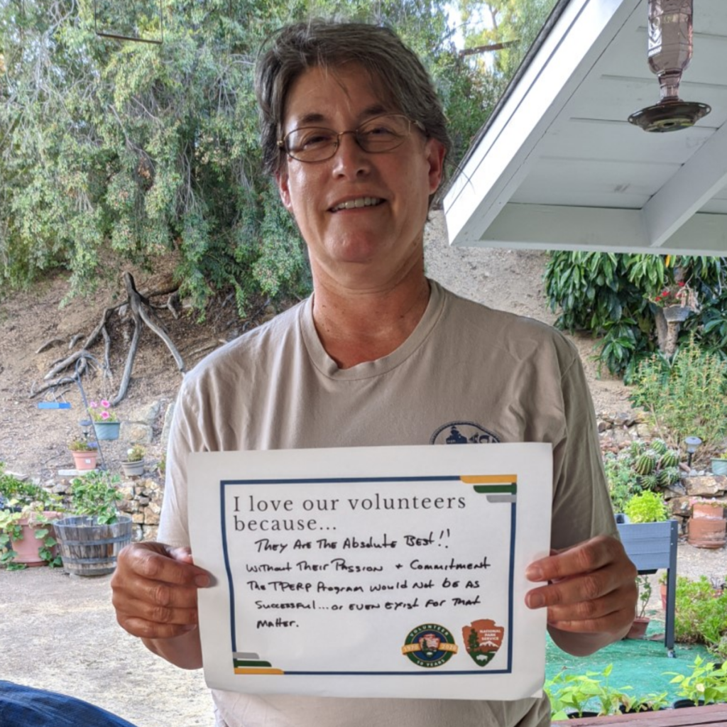 An up-close woman with a tan T-shirt smiles outside in front of a green and brown backyard and holds up a sign that says, “I love our volunteers because…they are the absolute best!! Without their passion and commitment, the TPERP [Tidepool Protection Education and Restoration] Program would not be as successful…or even exist for that matter.”