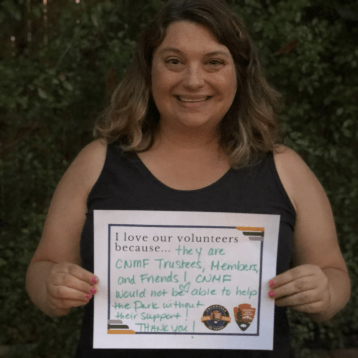 An up-close woman with a black blouse smiles outside in front of a dark green and leafy background and holds up a sign that says, “I love our volunteers because…they are CNMF [Cabrillo National Monument Foundation] trustees, members, and friends! CNMF would not be able to help the park without their support! Thank you!”