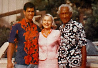 Miss Billy Riley in a pink dress standing between Kimo McVay wearing a colorful blue shirt with orange flowers and The Duke” Kahanomoku wearing a blue with white flowers Hawaiian shirt.