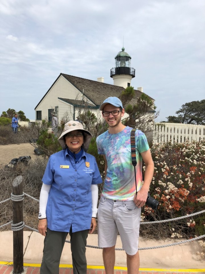 A woman in a blue shirt and sun hat stands next to a man in a tie dye shirt and blue baseball cap in front of a lighthouse.