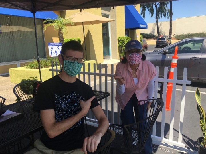 A man in a black shirt and face mask sits in a chair under an umbrella points to a woman wearing a face mask and a pink shirt.