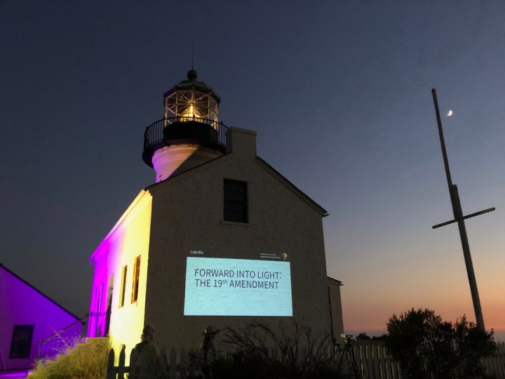 Lighthouse at night lit up with purple and gold with slideshow displayed on side of building. A flagpole with the moon above it is to the right of the building.