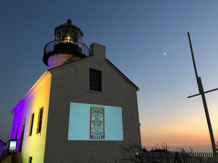Lighthouse at night lit up with purple and gold with slideshow displayed on side of building. A flagpole is to the right with the moon above it.