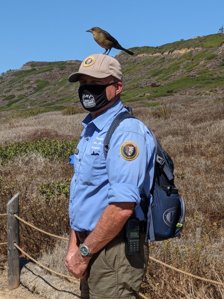 Man with black face mask and tan baseball hat and blue volunteer shirt with a brown and black bird on top of his hat.