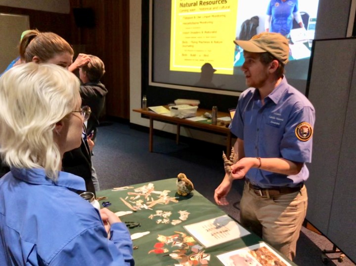 Park volunteer Wyler stands behind a green table in the park auditorium and holds a brown rubber mold of a bird’s feet and interacts with other park VIPs during a Science Explorer’s Club training. Another VIP and staff member interact separately and away from the camera, and a projector screen shows content about the Science Explorer’s club.
