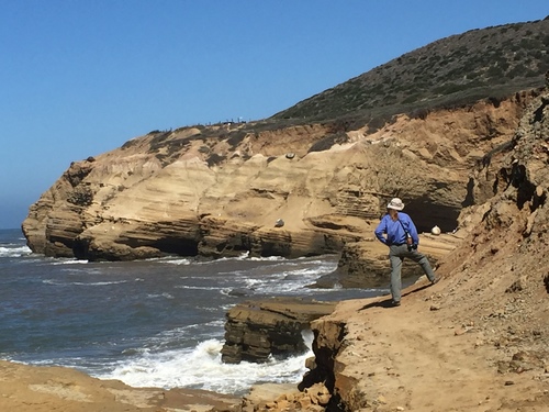 A shot from behind as a volunteer stands on a sandstone cliff at the tidepool area. The hillside turns upward sharply to her right. To the left the cliff drops off into the turbulent waters of the ocean.