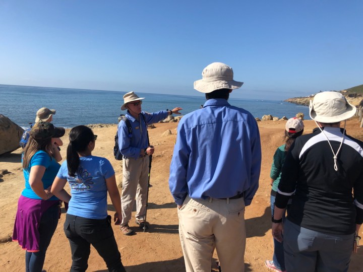 VIP Bob stands on a cliff near the tidepools while motioning to his left in front of a group of visitors and one other volunteer, who are all facing away from the camera.