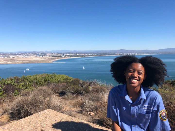VIP Felix sits on a seating area behind the Visitor Center and smiles at the camera. Behind her are various green-and-gray plants, the San Diego bay containing a few white sailboats and ships, the San Diego skyline, and mountain ranges, all under a blue cloudless sky.