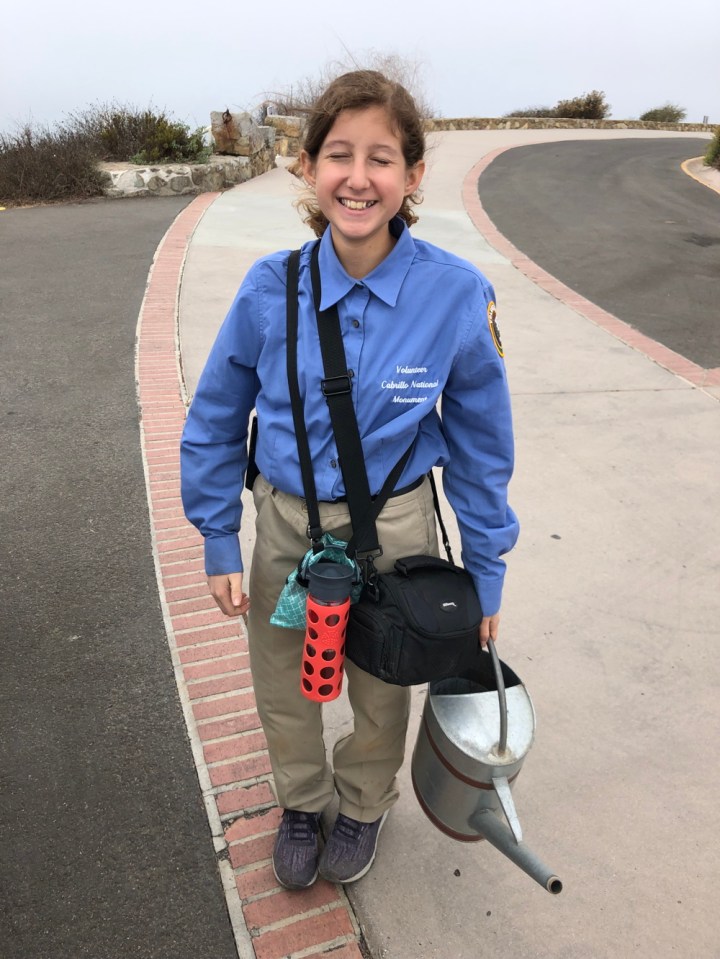 VIP Violette stands on a paved road behind the Old Point Loma Lighthouse with eyes closed and carrying a metal watering can, a photo camera, and a water bottle.