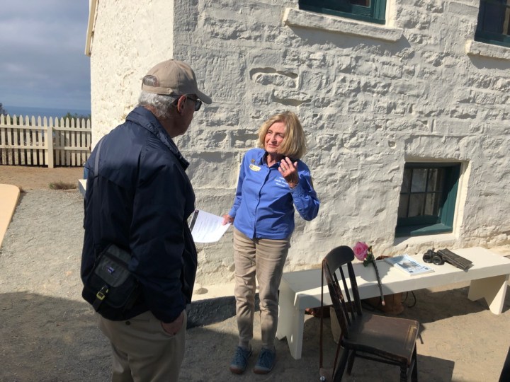 VIP Kim stands in front of the Old Point Loma Lighthouse, next to a bench and chair holding an assortment of props, while talking to another VIP not facing the camera during an Open Tower Day event.