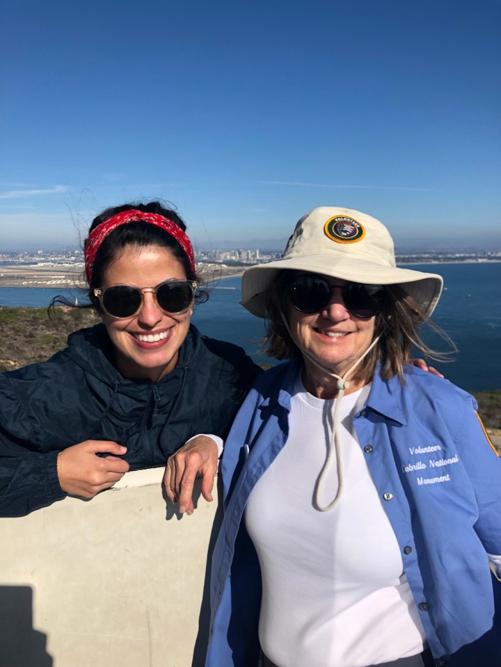 CVA Setareh, behind a short and white wall, and VIP Gayle stand on top of the Old Point Loma Lighthouse’s catwalk and smile at the camera. Behind them is a very small Cabrillo statue atop a hill, the San Diego bay and skyline, and mountain ranges.