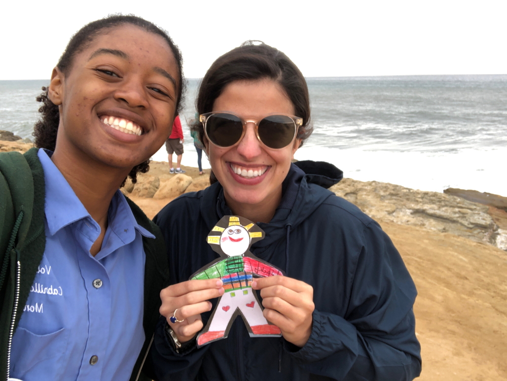 Park volunteer Felix wears an unzipped dark-green jacket and stands on a cliff next to CVA Setareh, who is wearing a zipped navy-blue windbreaker with sunglasses and holding a colorful Flat Stanley cutout. Both Felix and Setareh are smiling at the camera and facing away from the ocean. Two people stand at the edge of the cliffs behind Setareh and Felix, with their faces away from the camera.