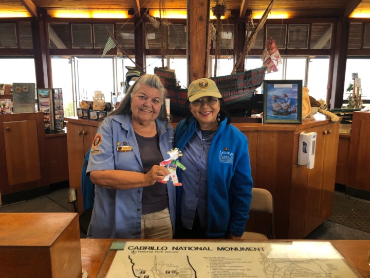 Smiling volunteers Barbara and Romi stand side-by-side behind a desk in the Visitor Center. They hold up a laminated, hand-drawn, colorful paper doll. Behind them is a replica of Cabrillo’s sailing ship, the San Salvador.