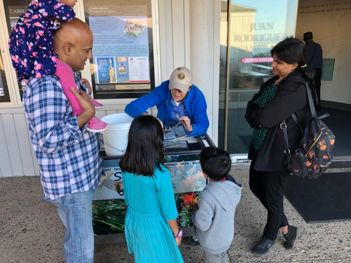 Volunteer Cecilia stands behind a rolling cart that’s labeled “Science Explorer’s Club”. On top of the cart is a white bucket, striped rubber snake, and reptile field guide that Cecilia uses to engage a family of two adults and three children.