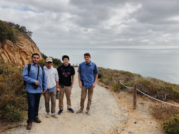 From left to right, park apprentices Alberto, Jackson, Christopher, and Eric stand side-by-side at the Bayside Trail and smile at the camera, with their backs to the ocean and overcast sky. Alberto and Eric wear park binoculars, and Jackson and Christopher wear hoodies that cover up their light-blue volunteer uniforms