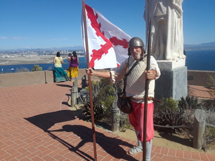 VIP Gary stands in front of the Cabrillo statue and holds a white flag with red lines in one hand and a spear in the other. He is dressed in 16th century Spanish soldier garb: a metal helmet, a thick white vest, short red pants, long gray socks, and tan woven sandals.