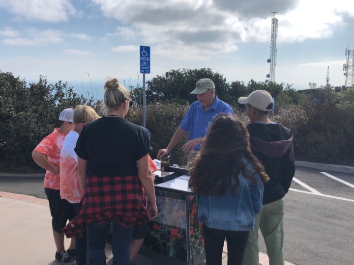Park volunteer Mike stands at an outdoor cart decorated with red flowers. Four children and one adult stand near him while he talks.