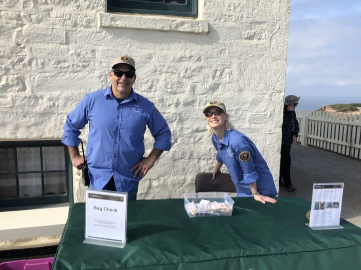 Volunteers Nick and Lizzie stand outside in front of a white building behind a green table with a sign that says Bag Check.