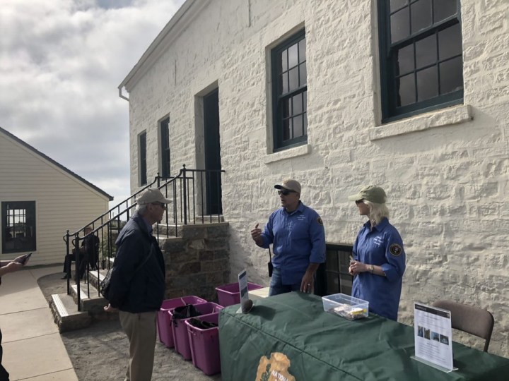 Volunteers Nick and Lizzie stand at green table with a Bag Check sign in front of a white brick building. Pink plastic boxes filled with backpacks are next to them. Nick talks to a park visitor wearing a bag over his shoulder.