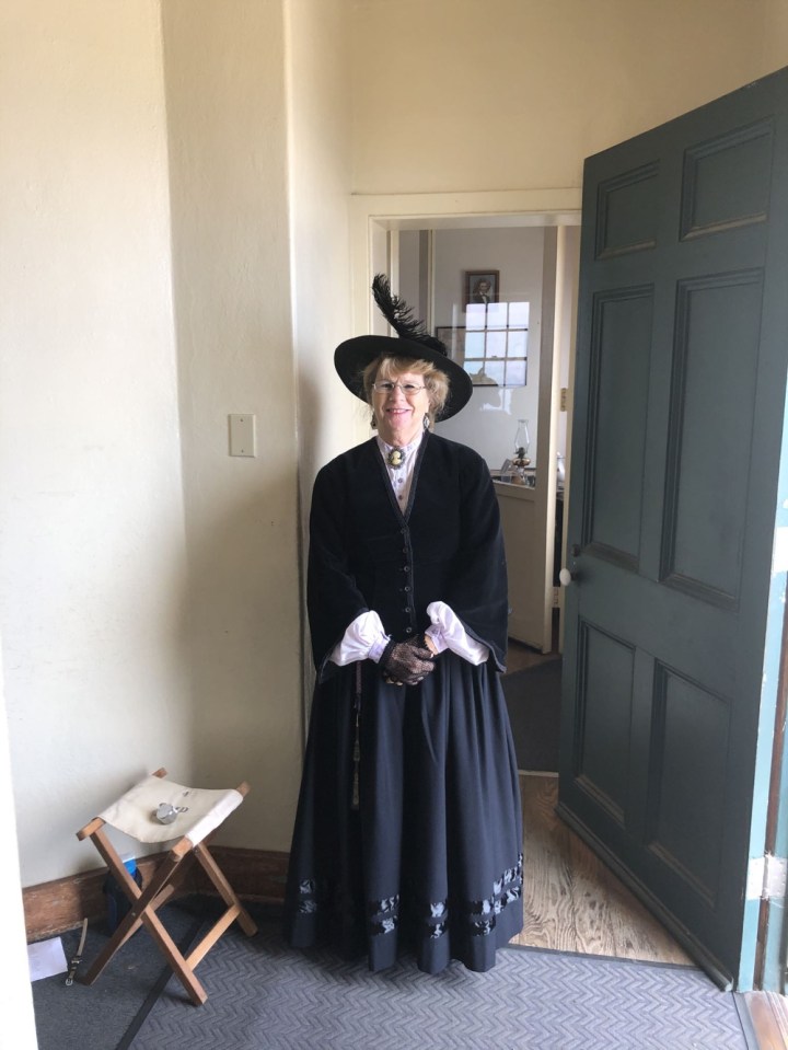 The large green front door of the lighthouse opens into the white foyer. Vicki, a volunteer stands inside the foyer ready to greet visitors wearing a dark blue dress and hat from the 19th century.