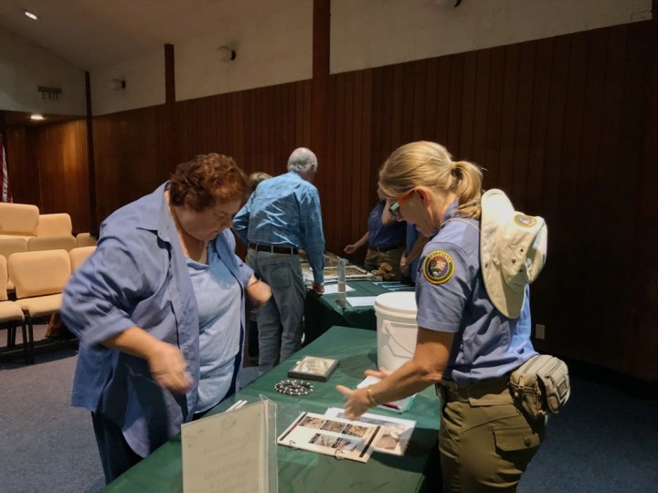 Park volunteer Katrina stands behind a green table in the park auditorium, points to the table, and interacts with park volunteer Jeannine during a Science Explorer’s Club training. A few VIPs stand further behind the table and interact separately.