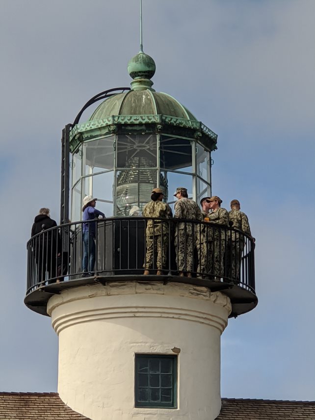 A view of the lighthouse tower. Five service members enjoy the view from the black iron lens deck, while volunteer Mayra stands by for questions. The Fresnel lens is visible in the center of the catwalk.