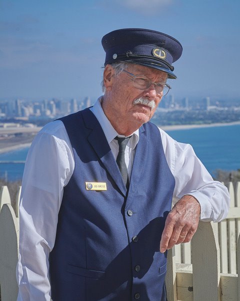 Volunteer Jack, wearing the dark blue vest and hat of a 19th century lighthouse keeper leans against the white picket fence of the rain catchment basin. The bay and San Diego are visible in the distance.