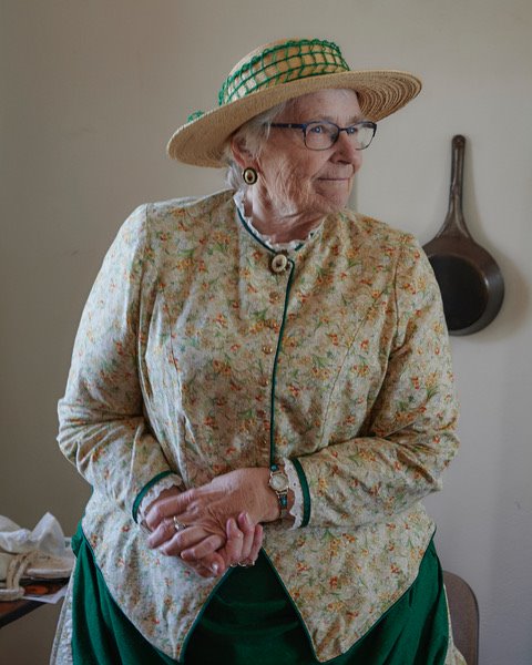 Volunteer Suzanne wearing a light yellow floral top and dark green dress in the style of the 19th century. Eileen is standing in the kitchen of the lighthouse with a large cast iron pan hanging from the wall behind her.