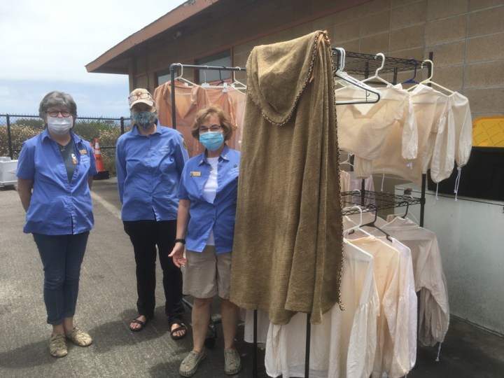 Volunteers Ferol, Dess, and Heide stand next to racks full of clothing outside in the lower maintenance area. The volunteers are all wearing masks.