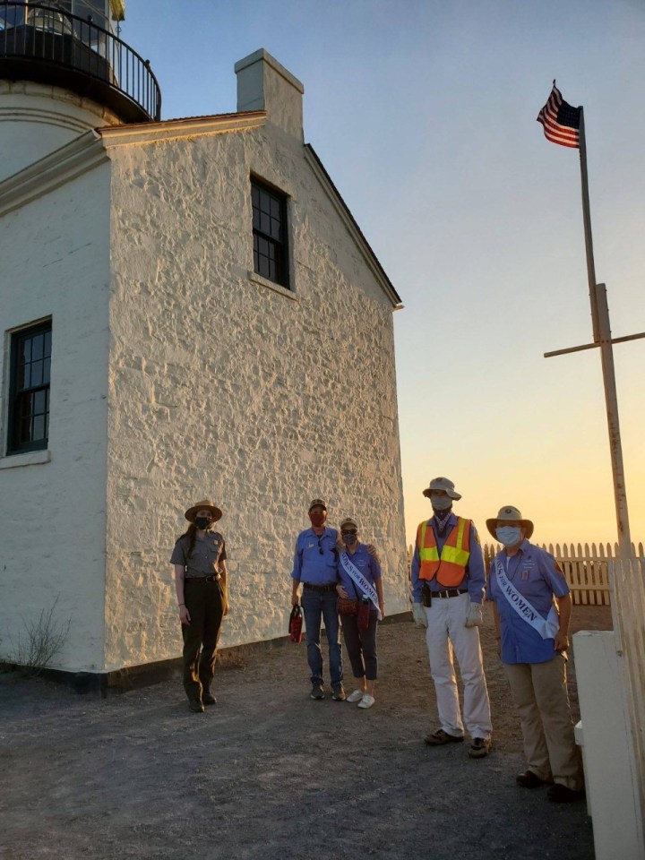 Park Ranger Julieanne, park volunteers Dan and Becky Wieder, Jose, and Sandy stand near the white brick wall of the lighthouse with the American flag behind them. All are wearing face masks. Becky and Sandy are wearing sashes across their uniforms that say Votes for Women.