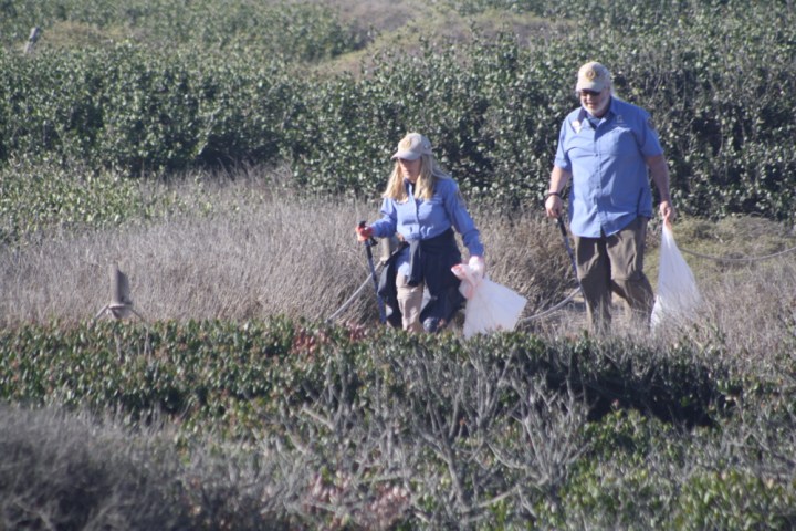 Park volunteers Carol and Larry hold a white trash bag in one hand and a long stick in the other hand. They walk along a rope fence surrounded by bushes.