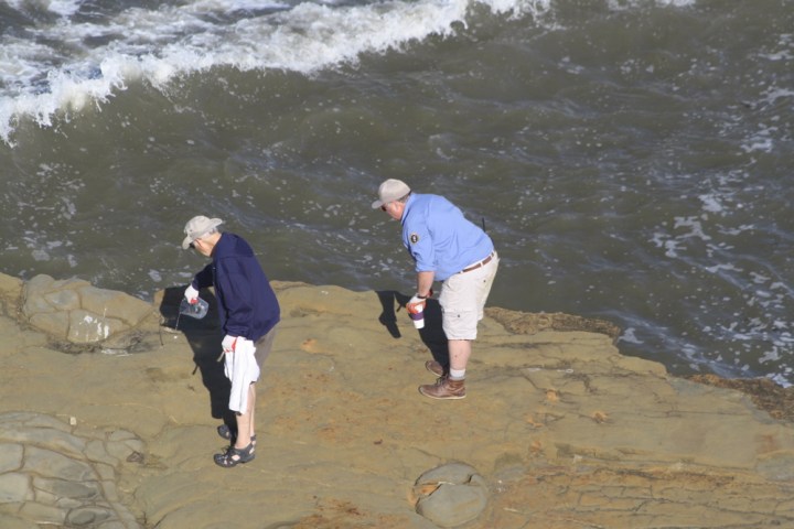 Volunteer Ken and Dave Stand on a rock in the ocean with a wave about to crash near them. Ken pours liquid from a jug on the ground.