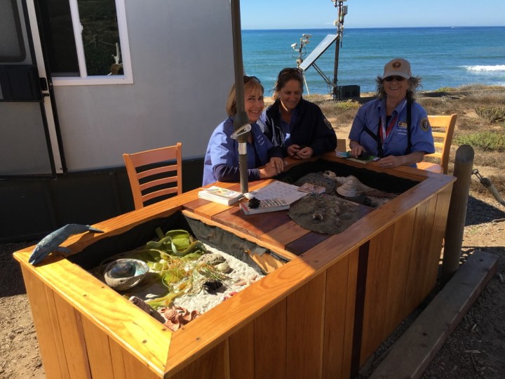 Tidepool VIPs, Pam, and former VIP Francie sit by tidepool education table with a handful of palm-sized science models and variety of science guides and spread out on the table in front of them. Tidepool trailer is to their left and view of coastal sage scrub and ocean is in the distance to their right.
