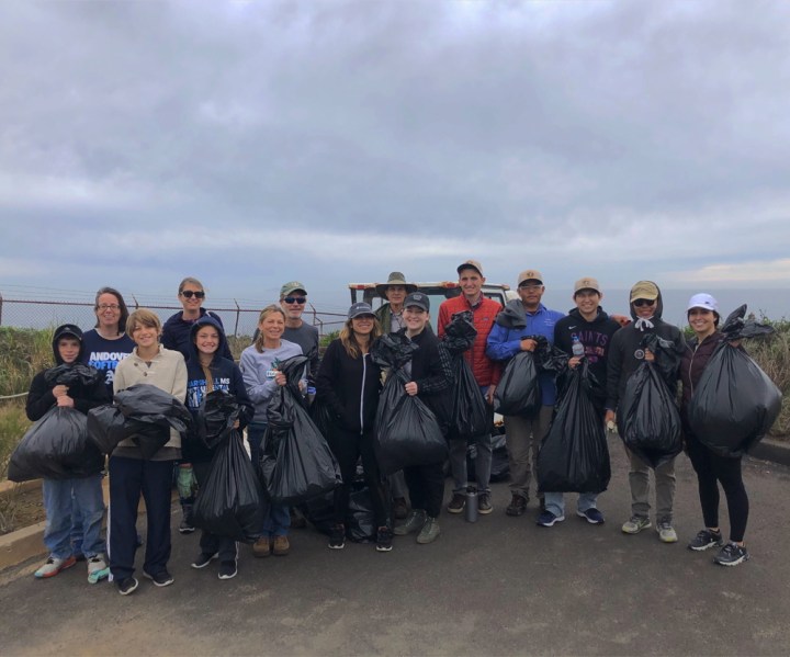A group of fifteen people stand together on a paved path, holding ten, half-way filled trash bags. Above them is an overcast sky and behind them are roped-off green hedges and a walkway, in which three people on the far-right walk on.