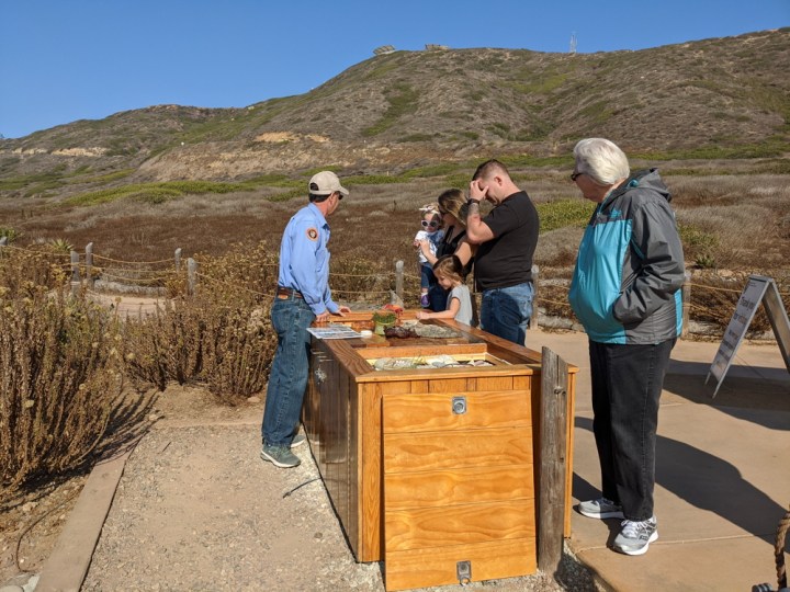 Park volunteer Dan stands behind the tidepool Education Table talking to two children and three adults about what they can find in the tidepools. The cement portion of the Coastal Trail is in the background along with brown and green bushes.