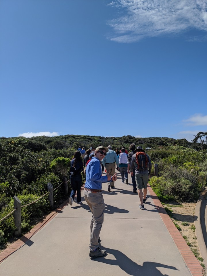 A view from behind as a large group of volunteers and staff head up the path that leads to the lighthouse. Apprentice Wyler, who is bringing up the rear of the group, turns to face us with a smile and a thumbs up.