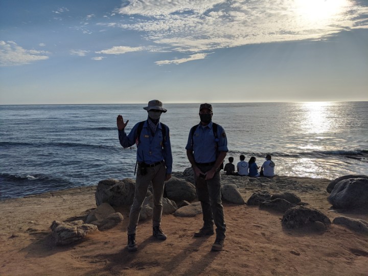 VIPs Jose & Brian stand and pose on a sandy cliff in front of sunset over the water. 4 visitors sit on the edge of the cliff behind them and look out at the water.