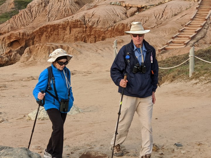 VIPs Elke & Bob walk along a dirt path near steps on a sandstone cliff. They wear a jacket, sunglasses, binoculars, and use a hiking stick.