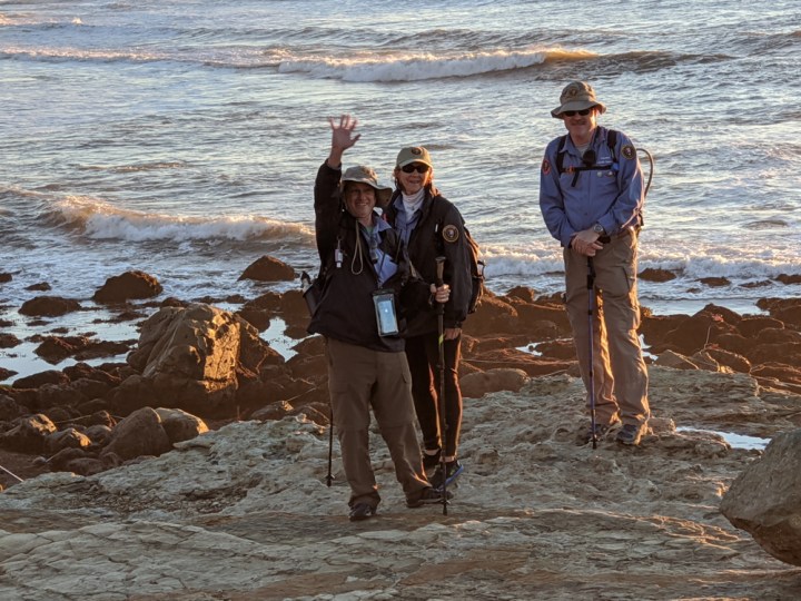 VIPs Dan, Mary, & Sean stand and pose on an uneven, dry path in front of small, brown boulders, and the vast ocean with white-topped waves.