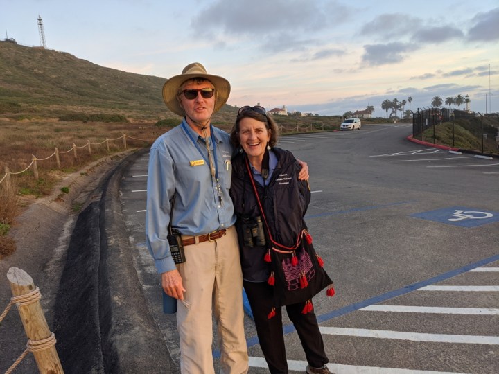 VIP Bob wraps his arm around VIP Julie while they stand & pose in a parking area just west of a grassy hill.