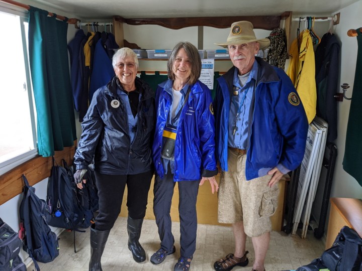 Tidepool VIPs Eileen, Gayle, and Patrick smiling and wearing blue windbreakers in the tidepool trailer. Eileen is wearing rubber boots while Gayle and Patrick wear closed-toed water sandals.