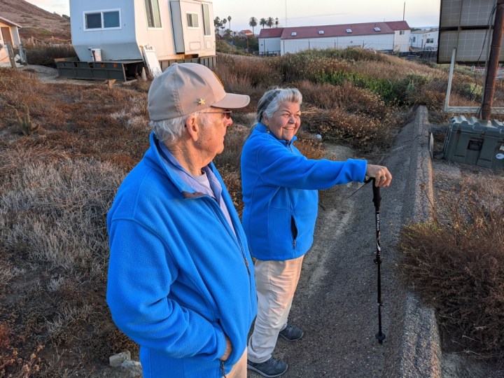 Tidepool VIPs Barbara and Ken in blue fleece VIP jackets stand by concrete embankment with tidepool trailer and Navy buildings behind them. Barbara is smiling looking over the horizon and holds a walking stick. Ken’s profile is visible as he looks away from the camera and to the horizon in the other direction.