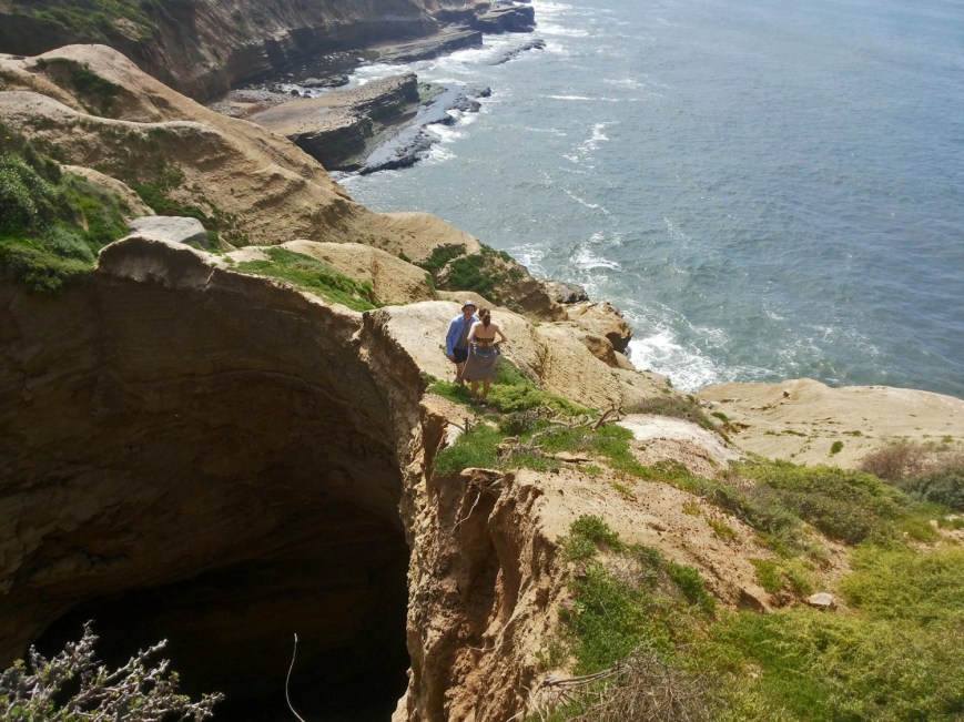 Two people stand at a dangerous edge of a sinkhole along sandstone cliffs at the ocean's edge.