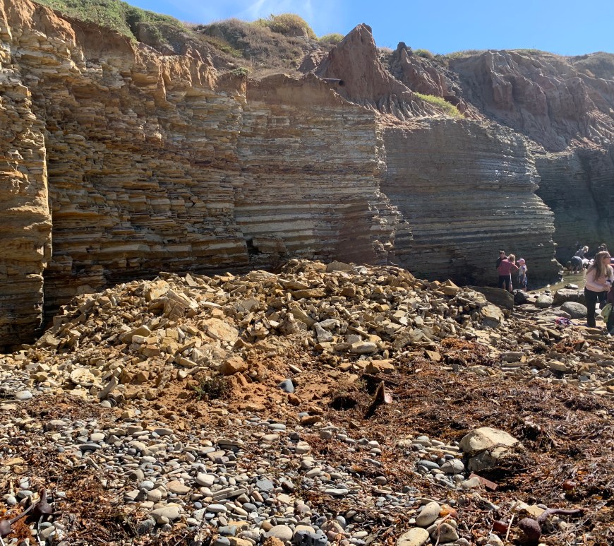 Broken sandstone rocks against a layered cliff. Several people are in the background looking at the rock fragments.