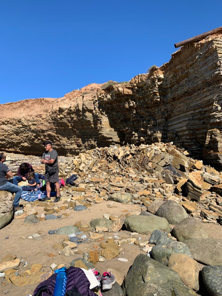 Broken sandstone rocks against a layered cliff. Several people are in front of the chunks of rock.